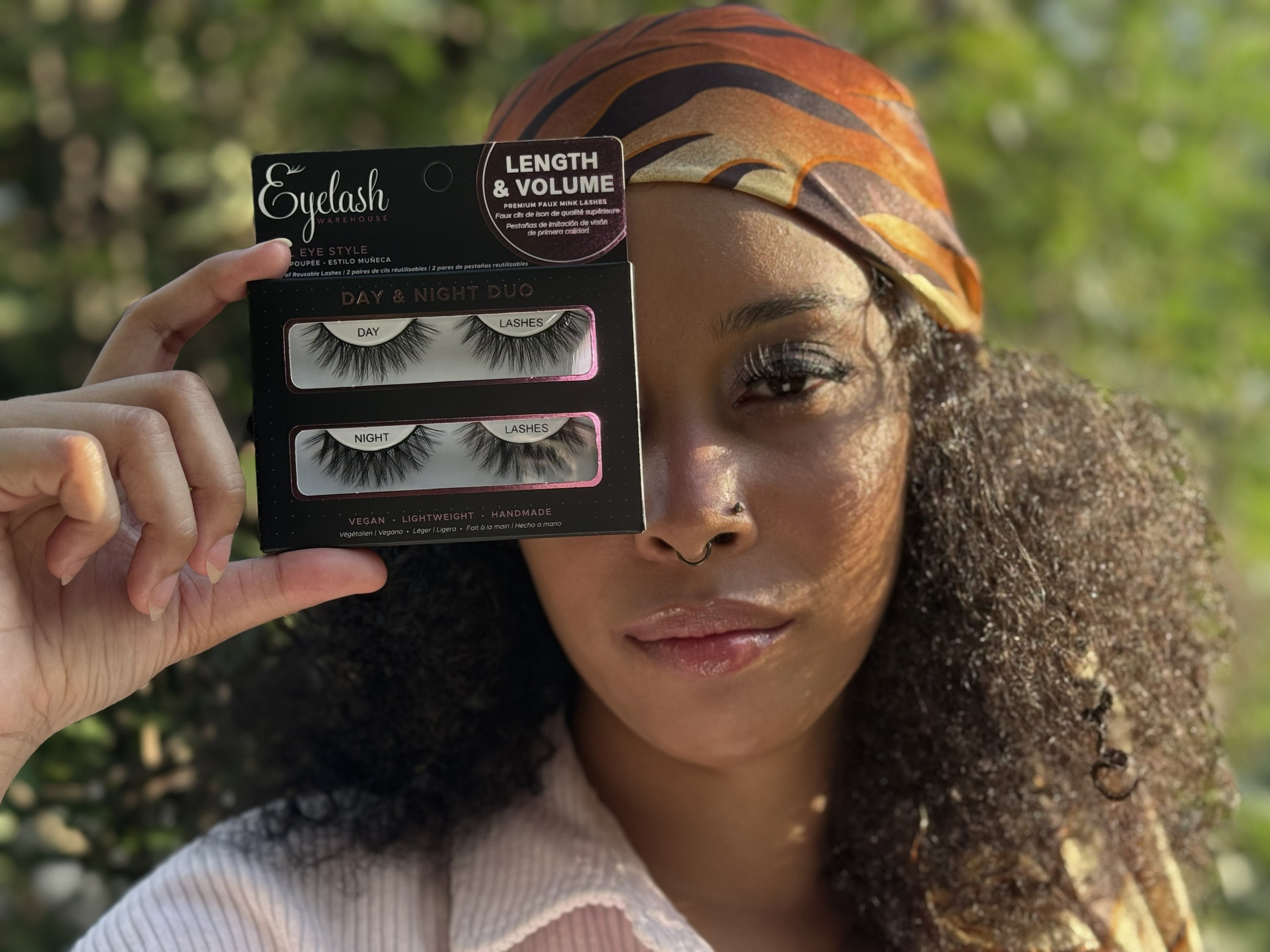 A woman holding a pair of false eyelash packages in front of her face, with a blurred outdoor green background. The packages are labeled 'Eyelash' and 'Day & Night Duo'.